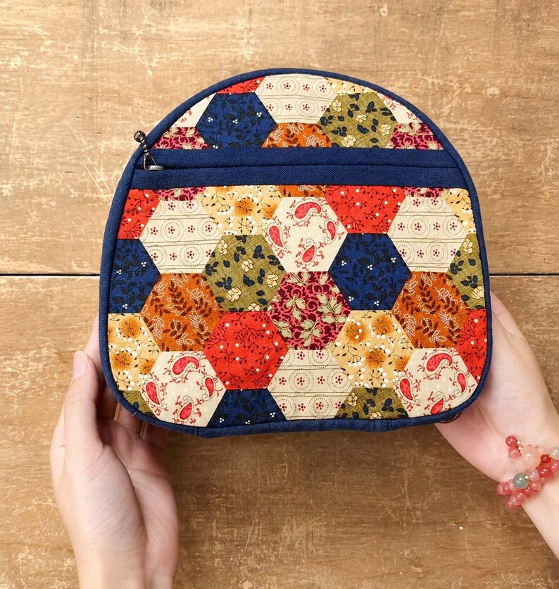 May include: A semi-circular zipped pouch featuring a patchwork design of multi-coloured fabric hexagons. The exterior displays various floral and paisley patterns in shades of red, navy blue, orange, and cream, finished with a solid navy blue trim.
