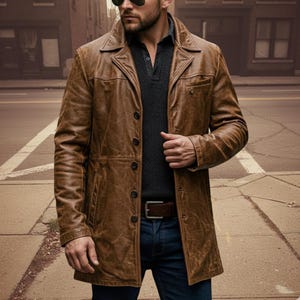 May include: A brown leather jacket with a button-up front, a notched collar, and a belt. The jacket is worn over a dark gray sweater and blue jeans. The jacket appears to be made of distressed leather.