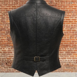K&ouml;nnte beinhalten: Schwarze Lederweste mit einem Reverskragen und einem Schnallenverschluss an der Taille. Die R&uuml;ckseite der Weste hat ein gen&auml;htes Design und ein kleines Kreuzstichdetail in der N&auml;he des Kragens. Die Weste wird vor einer Backsteinmauer gezeigt.