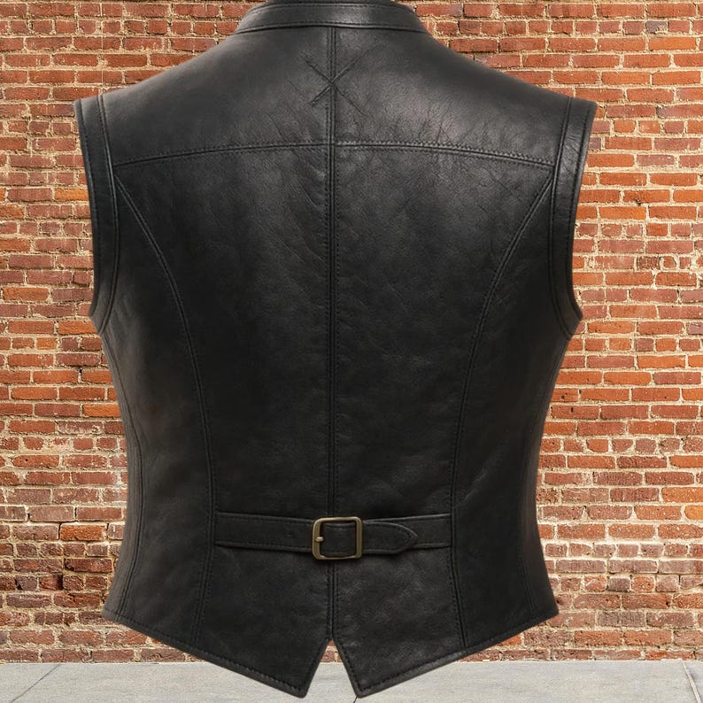 K&ouml;nnte beinhalten: Schwarze Lederweste mit einem Reverskragen und einem Schnallenverschluss an der Taille. Die R&uuml;ckseite der Weste hat ein gen&auml;htes Design und ein kleines Kreuzstichdetail in der N&auml;he des Kragens. Die Weste wird vor einer Backsteinmauer gezeigt.