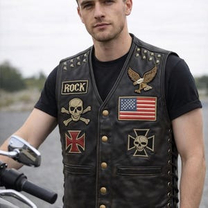 Könnte beinhalten: Eine schwarze Lederweste mit verschiedenen Aufnähern, darunter ein Totenkopf, eine amerikanische Flagge und das Wort "ROCK". Die Weste hat goldfarbene Knöpfe und Nieten an den Schultern. Getragen über einem schwarzen T-Shirt.