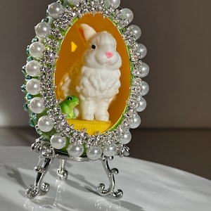 May include: An Easter-themed decorative egg with a white bunny and green frog inside. The egg is surrounded by faux pearls and rhinestones, and sits on a silver stand. The egg's interior is yellow and green.