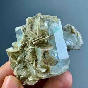 May include: A raw aquamarine crystal cluster with pale blue and clear sections. The crystal has a rectangular shape with a light green hue. Surrounding the crystal are clusters of beige mica.