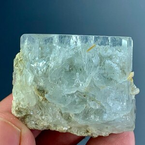 May include: A raw aquamarine crystal specimen. The crystal is a pale blue-green color with a rectangular shape and a rough, textured surface. The base of the crystal is a light tan color. The crystal is held in a hand.