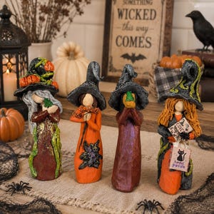 May include: Four standing witch figurines with pointed hats, each wearing long dresses in shades of orange, brown, and black. The figures hold various items including a small green frog, a bouquet of flowers, and a bag labeled Hocus Pocus.
