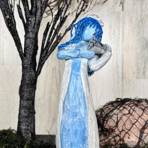 May include: A carved wooden figurine of a blue-skinned woman in a white and blue dress, holding a gray object. The figure has blue hair and is standing in front of a black tree and green moss.