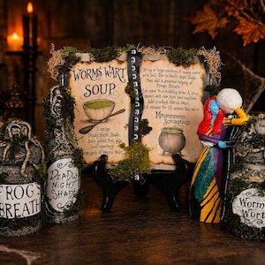 May include: A decorative display featuring a central sign titled Worms Wart Soup with a recipe, flanked by three textured jars labeled Frog's Breath, Deadly Night Shade, and Worm's Wort, all adorned with moss, alongside a figurine of two characters.