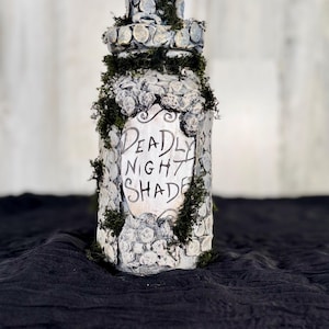 May include: A decorative bottle with the words "Deadly Night Shade" in black script. The bottle is white with a stone-like texture and is adorned with green moss. The bottle has a matching stone-like cap.
