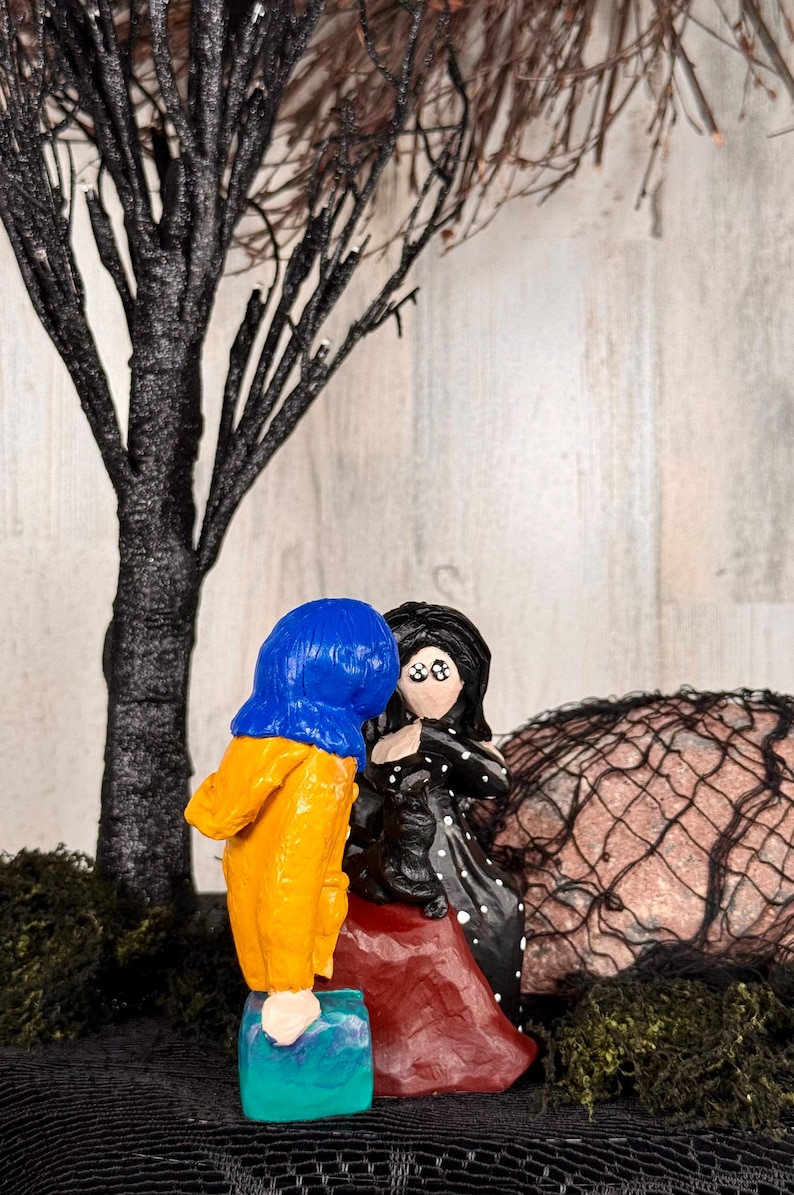May include: A handcrafted figurine set featuring two figures, one in a blue and yellow outfit, the other in a black dress with white polka dots. The figures are positioned near a black tree and a decorative net.