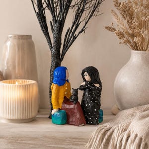 May include: A decorative figurine set featuring two figures, one in a yellow jacket and blue hair, the other in a black polka dot dress. A black cat sits between them. The figures are seated on turquoise blocks, with a black tree and vase in the background.