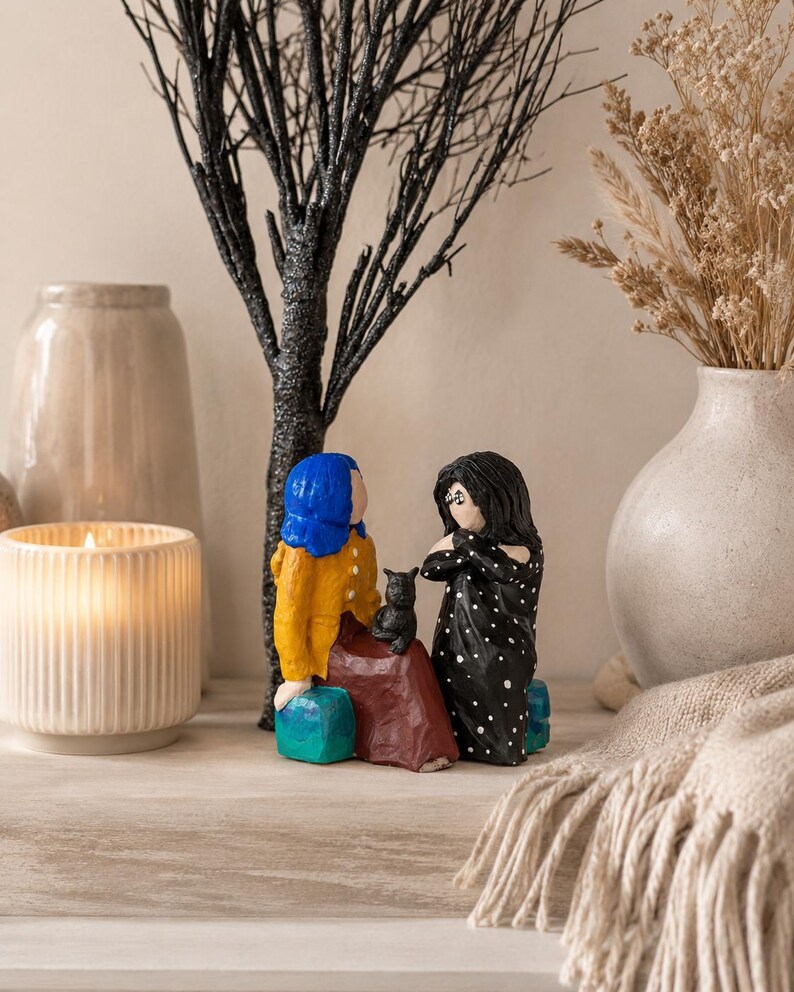 May include: A decorative figurine set featuring two figures, one in a yellow jacket and blue hair, the other in a black polka dot dress. A black cat sits between them. The figures are seated on turquoise blocks, with a black tree and vase in the background.