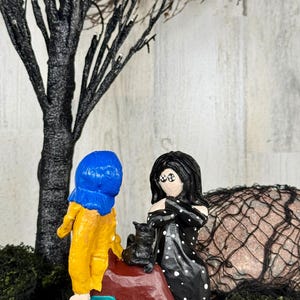 May include: A whimsical diorama featuring two stylized figures, one in a yellow coat and blue hood, the other in a black dress with white polka dots. A black cat sits on a red rock. A black tree and moss complete the scene.