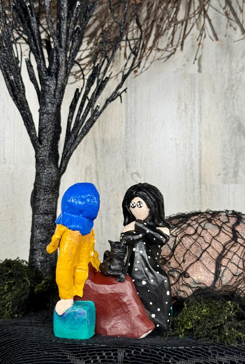 May include: A whimsical diorama featuring two stylized figures, one in a yellow coat and blue hood, the other in a black dress with white polka dots. A black cat sits on a red rock. A black tree and moss complete the scene.