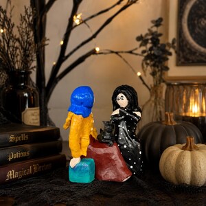 May include: A whimsical Halloween-themed display featuring handmade figurines. One figurine wears a blue hat and yellow outfit, another a black dress with white polka dots. A black cat sits atop a red cape. Decorative pumpkins and books with titles like "Spells" and "Potions" complete the scene.