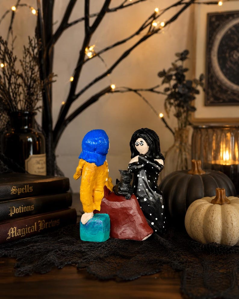 May include: A whimsical Halloween-themed display featuring handmade figurines. One figurine wears a blue hat and yellow outfit, another a black dress with white polka dots. A black cat sits atop a red cape. Decorative pumpkins and books with titles like "Spells" and "Potions" complete the scene.