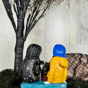 May include: A handcrafted figurine depicting two figures seated together. One figure wears a black coat with white polka dots, the other a yellow raincoat and blue hood. They sit on a blue base, with a black tree and moss in the background.