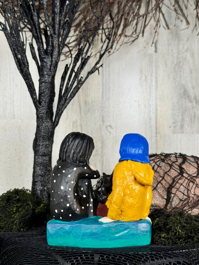 May include: A handcrafted figurine depicting two figures seated together. One figure wears a black coat with white polka dots, the other a yellow raincoat and blue hood. They sit on a blue base, with a black tree and moss in the background.