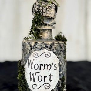May include: A decorative bottle with a textured, gray and gold finish, adorned with green moss. The label reads "Worm's Wort" in elegant script. The bottle's design has a vintage, apothecary aesthetic.