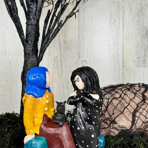 May include: A whimsical, handcrafted figurine set featuring two figures and a black cat. One figure wears a blue wig and yellow coat, the other a black dress with white polka dots. A black tree and decorative moss complete the scene.