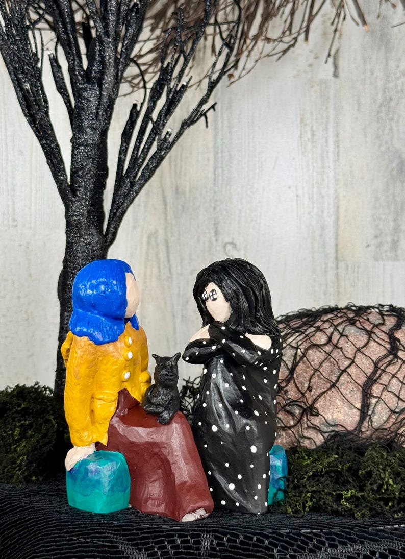 May include: A whimsical, handcrafted figurine set featuring two figures and a black cat. One figure wears a blue wig and yellow coat, the other a black dress with white polka dots. A black tree and decorative moss complete the scene.