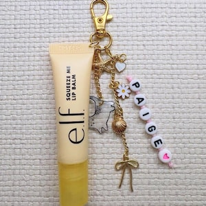 May include: A tube of e.l.f. Squeeze Me lip balm with a gold-coloured keychain. The keychain has charms including a star, heart, daisy, seashell, and a bow. A beaded strand spells out the name "Paige".