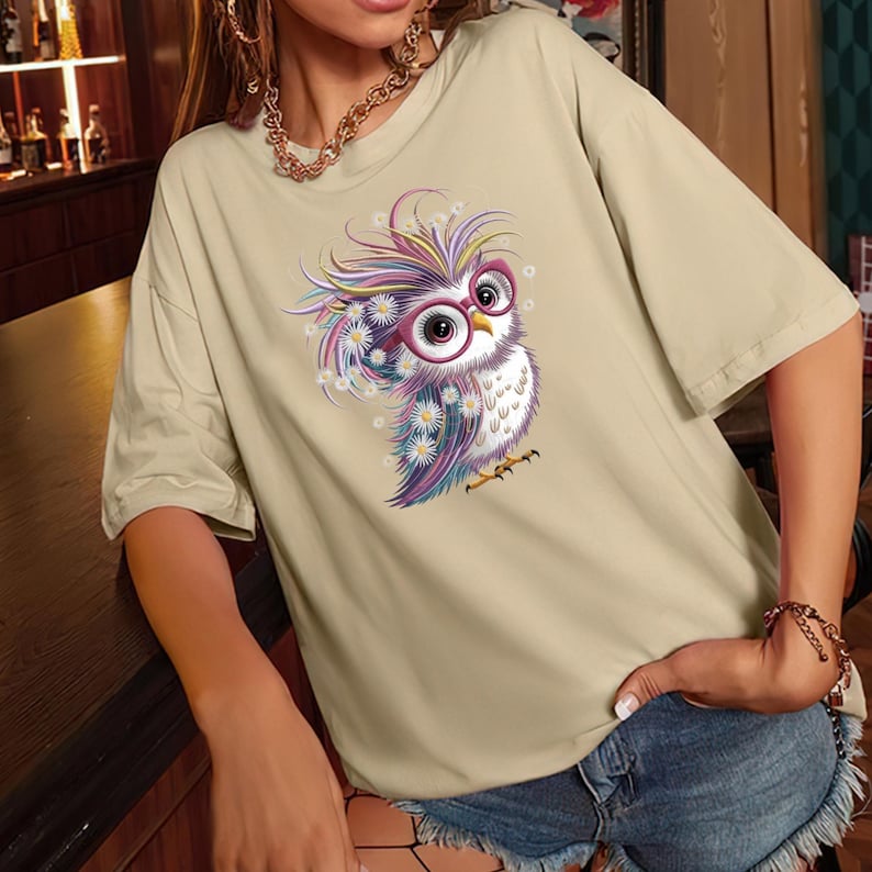 May include: Beige short-sleeved t-shirt featuring a colorful cartoon owl wearing glasses. The owl has a white chest, purple and blue feathers, and is surrounded by white flowers. The shirt is made of a soft material.