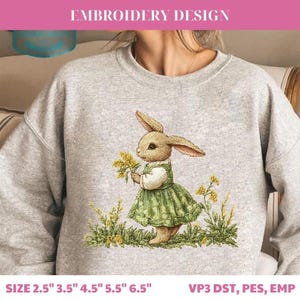 May include: A gray sweatshirt featuring an embroidered design of a bunny wearing a green dress and holding yellow flowers. The text "EMBROIDERY DESIGN" is at the top. Below the design are the sizes 2.5", 3.5", 4.5", 5.5", and 6.5".