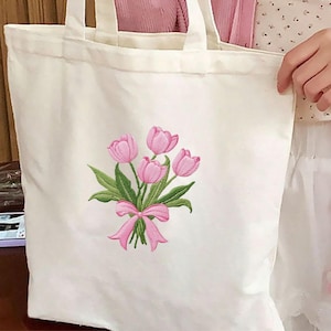 May include: A white canvas tote bag featuring an embroidered bouquet of pink tulips with green leaves and a pink ribbon. The tote bag has two white handles.