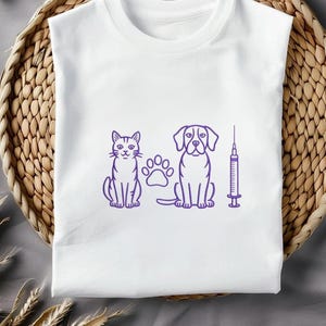 May include: White t-shirt featuring a purple line drawing of a cat, a dog, a paw print, and a syringe. The cat and dog are sitting upright. The design is centered on the shirt.
