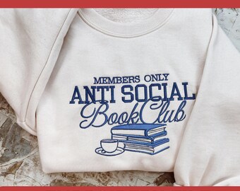 May include: A cream-coloured sweatshirt featuring blue embroidered text: "MEMBERS ONLY ANTI SOCIAL Book Club." Below, an embroidered image of books and a teacup. The text "EMBROIDERY FILE" is at the bottom. The size is 10, 12, 15, 18, and 20 cm.