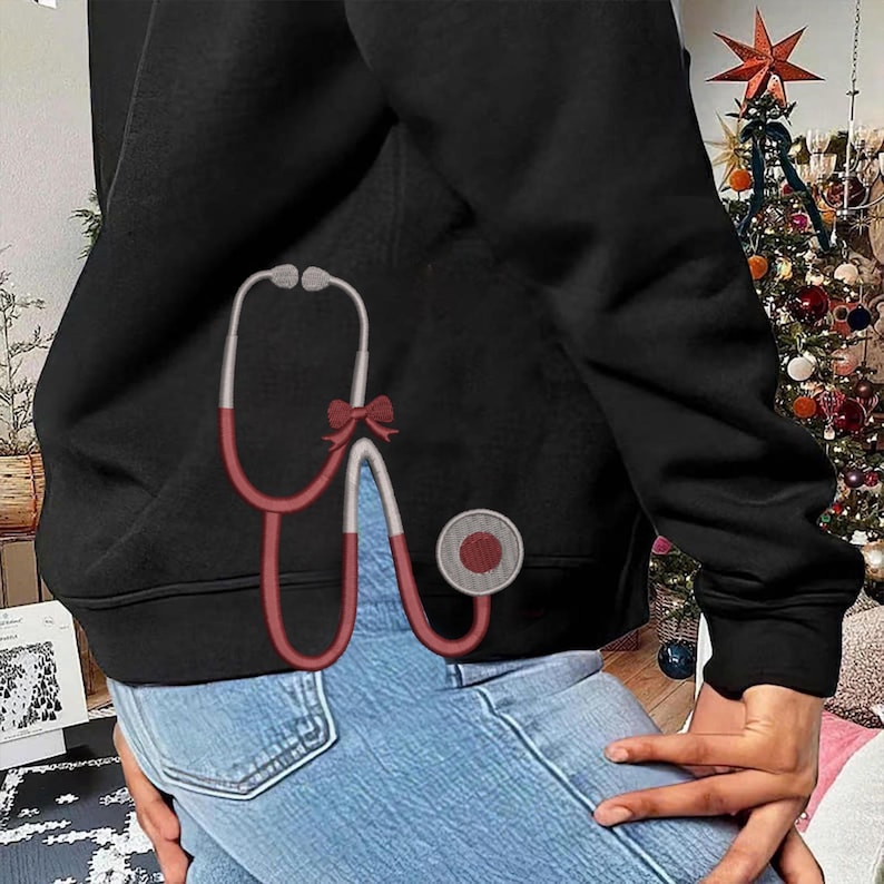 May include: Black hoodie with a red and white stethoscope design. The stethoscope has a red bow and a gray circular piece. The hoodie is paired with blue jeans. A Christmas tree is visible in the background.