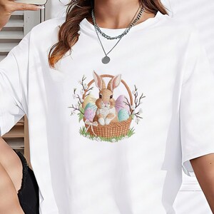 May include: White t-shirt featuring an embroidered Easter design. The design includes a brown bunny sitting in a woven basket with colorful Easter eggs. The basket is surrounded by greenery and branches with white flowers.