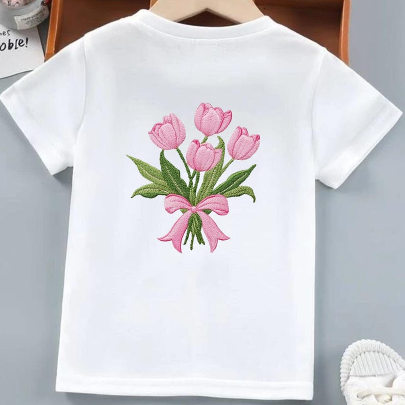 May include: White t-shirt featuring an embroidered bouquet of pink tulips with green leaves and a pink bow. The short-sleeved shirt is made of a soft material and is suitable for children's wear.
