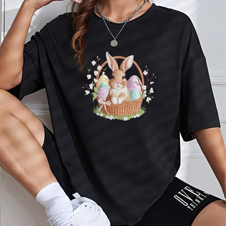 May include: Black t-shirt featuring an Easter bunny in a basket with colorful eggs and flowers. The graphic is centered on the front of the shirt. The shirt is a casual, short-sleeved style.