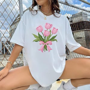 May include: White t-shirt featuring an embroidered bouquet of pink tulips tied with a pink bow. The floral design includes green leaves. The shirt has short sleeves and a relaxed fit, suitable for casual wear.