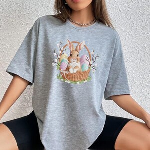 May include: A gray t-shirt featuring an Easter-themed graphic. The design shows a brown bunny sitting in a woven basket filled with colorful Easter eggs. The basket is surrounded by sprigs of white flowers and green grass.