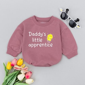May include: A mauve-colored baby bodysuit with the text "Daddy's little apprentice" and a lightbulb graphic. The long-sleeved outfit has a round neckline and ribbed cuffs. Sunglasses shaped like a giraffe are placed on the side.