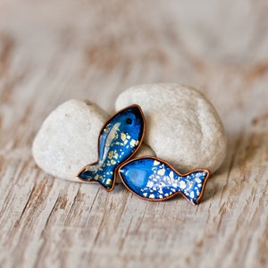 Puede incluir: Dos pendientes de resina en forma de pez azul y dorado con detalles en cobre. Los pendientes están sobre un fondo blanco.