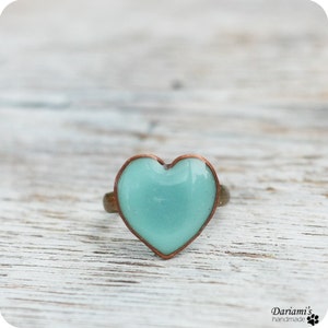 K&ouml;nnte beinhalten: Ein herzf&ouml;rmiger Ring mit hellblauem Zentrum und kupferfarbenem Rand. Der Ring hat ein bronzefarbenes Band. Der Ring befindet sich auf einer wei&szlig;en Holzoberfl&auml;che. Der Ring ist handgefertigt.