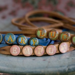 May include: Two leather bracelets with colourful glass beads spelling out the names "MARY" and "EMILY". The beads are blue, pink and yellow.