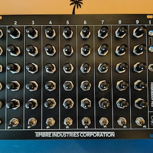 May include: A black panel with 64 toggle switches arranged in an 8x8 grid. The panel is labelled "TIMBRE INDUSTRIES CORPORATION" and "MAKER BUDDY MIDI".
