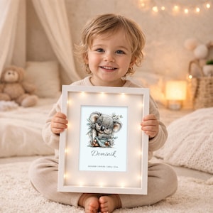 Könnte beinhalten: Ein weißer Bilderrahmen mit einer Aquarellillustration eines Koala-Bären. Der Rahmen hat kleine Lichter am Rand. Der Name "Dominik" steht unter dem Koala. Das Kind hält den Rahmen.