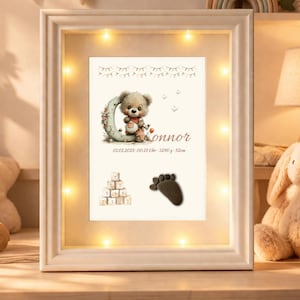Könnte beinhalten: Gerahmte Baby-Ankündigung mit Teddybär-Illustration, dem Namen "Connor" und Geburtsdetails. Der weiße Rahmen ist mit warmweißen Lichtern versehen. Enthält einen Babyfußabdruck und Holzblöcke.