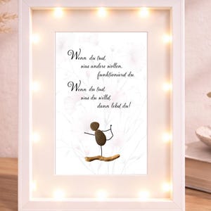 Könnte beinhalten: Ein gerahmtes Kunstwerk mit weißem Rand und kleinen Lichtern. Das Kunstwerk zeigt eine Strichmännchenfigur, die auf einem Holzstück balanciert, mit Text auf Deutsch. Der Hintergrund hat ein florales Design. Der Rahmen ist rechteckig.