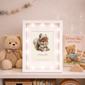 Könnte beinhalten: Ein weißer Bilderrahmen mit Lichterkette, der eine Aquarellillustration eines Eichhörnchens in Winterkleidung zeigt. Der Rahmen enthält den Namen "Annelie" und Geburtsdaten. Ein Teddybär und ein Buch sind ebenfalls zu sehen.