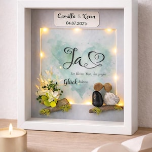 Könnte beinhalten: Ein gerahmtes Hochzeitsandenken mit den Namen "Camilla & Kevin" und dem Datum "04.07.2025". Das Kunstwerk zeigt ein Aquarellherz, das Wort "Ja" und ein Kieselkunstpaar. Dekorative Lichter und florale Akzente vervollständigen das Design.