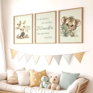 Könnte beinhalten: Drei gerahmte Kunstdrucke mit Aquarellillustrationen einer Giraffe, eines Tigerbabys und inspirierendem Text. Die Drucke befinden sich in hellen Holzrahmen und werden über einem Sofa mit dekorativen Kissen und Stofftieren ausgestellt. Über dem Sofa hängt ein Banner aus Dreiecksflaggen.