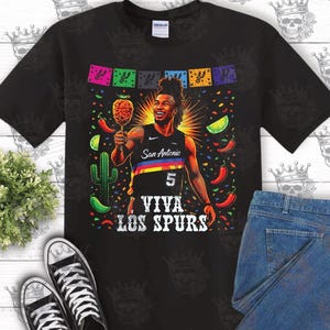 May include: Black t-shirt featuring a vibrant graphic of a basketball player holding a fried food item, with the text "San Antonio" and "Viva Los Spurs." The design includes colorful confetti, peppers, and a cactus.