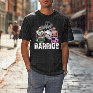 May include: Black t-shirt featuring a graphic of two boxers and the word "BARRIOS" in white block letters. The shirt has a crew neck and short sleeves. The boxers are wearing boxing gloves and trunks.