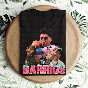 May include: Black t-shirt featuring a collage of images and the word "BARRIOS" in a pink and orange design. The shirt showcases multiple figures, including a boxer with pink gloves and other individuals. The design is printed on a black background.